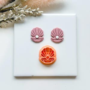 May include: Three seashell-themed items. Two pink seashell earrings with white pearl accents and a coral-coloured seashell-shaped cookie cutter. The items are arranged on a white surface with a sprig of dried flowers in the background.