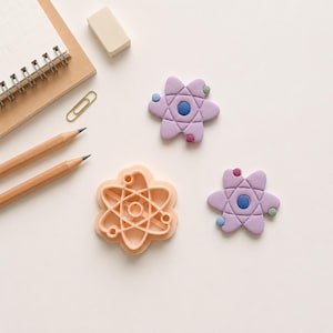 May include: Two lavender atom-shaped clay pieces with small blue, pink, and green circular accents, alongside a peach-colored plastic atom-shaped cookie cutter. The atom designs feature a central blue circle surrounded by petal-like shapes.