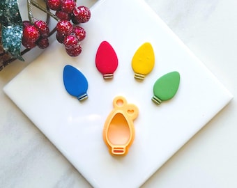 May include: Five colorful light bulb shaped cookie cutters on a white surface. The cutters are red, blue, yellow, green, and orange. The orange cutter has a heart-shaped detail.