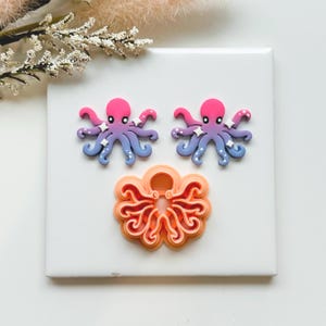 May include: Two octopus-shaped earrings and a cookie cutter. The earrings are pink, purple, and blue with white star accents. The cookie cutter is orange and has a detailed octopus design. All items are on a white surface.