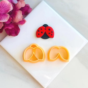 May include: Two cookie cutters in the shape of flower petals, and a red and black ladybug cookie cutter on a white surface.