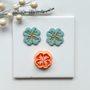 May include: Three clover-shaped craft items. Two are teal with gold accents, and one is orange. The items are arranged on a white surface, with a decorative pearl and silver branch in the background. The clover shapes are likely for crafting or jewelry making.