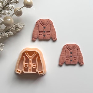 May include: A peach-coloured plastic cookie cutter shaped like a V-neck cardigan with two pockets, accompanied by two finished, textured, peach-coloured cardigan-shaped biscuits featuring dark grey buttons, arranged on a plain white surface.