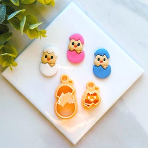 May include: A set of five colorful chick-themed polymer clay charms. The charms are in the shape of chicks hatching from eggs. The colors include white, pink, blue, and yellow.