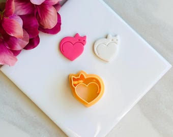 May include: Three cat-shaped heart cookie cutters in pink, white, and orange. The pink and white hearts are solid, while the orange one is a cutter. A pink flower is in the upper left corner. The objects are on a white surface.