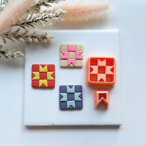 May include: An overhead shot displays a collection of craft supplies. There are three square, textured, colorful clay pieces with a starburst design, and two orange plastic cutters. The clay pieces are red, tan, and blue. The cutters are square and rectangular.