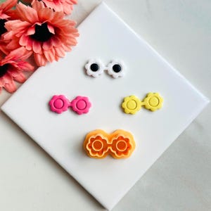 May include: Four cookie cutters in the shape of glasses with flower details. The cutters are white, pink, yellow, and orange.