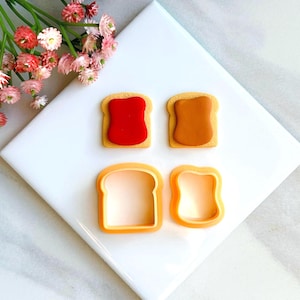May include: Two cookie cutters shaped like bread slices, one with peanut butter and one with jelly, and two finished cookies, one with peanut butter and one with jelly.