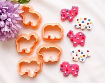 May include: A collection of five orange plastic animal-shaped cookie cutters alongside five corresponding animal-shaped cookies decorated with small, colorful dots in red, blue, and yellow. A large purple flower is positioned in the upper left corner.