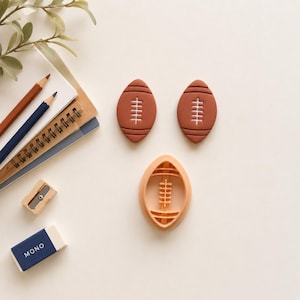 May include: Two solid brown football-shaped cookie cutters with white lace details and one light peach-colored football-shaped cookie cutter frame. The items are arranged alongside two colored pencils, a metal sharpener, and a blue eraser.