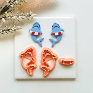 May include: A selection of shark-themed craft items. Two blue shark figurines with red and white life rings, and two sets of shark-shaped cookie cutters in shades of orange and pink. A small orange and pink cutter is also included.