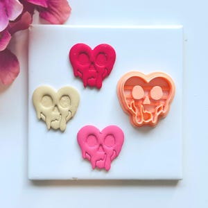 May include: Four heart-shaped objects with skull designs. One is a cookie cutter, and the others are finished products in shades of pink and cream. The designs have a dripping effect, and the objects are on a white surface.