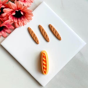 Puede incluir: Cuatro panecillos marrones en miniatura y un panecillo naranja con un diseño decorativo. Los panecillos están sobre una superficie blanca.