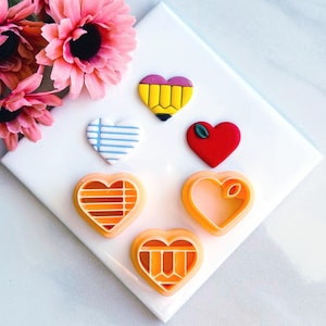 May include: Five heart-shaped cookie cutters, two are orange with a grid pattern, one is white with blue stripes, one is red with a green leaf, and one is pink with a yellow pencil design.