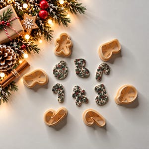 Może przedstawiać: Zestaw beżowych plastikowych foremek do ciastek w kształcie ludzików z piernika, skarpet, lasek cukrowych i ozdób, wraz z pasującymi ciasteczkami ozdobionymi wzorem zielonych liści ostrokrzewu i czerwonych jagód.