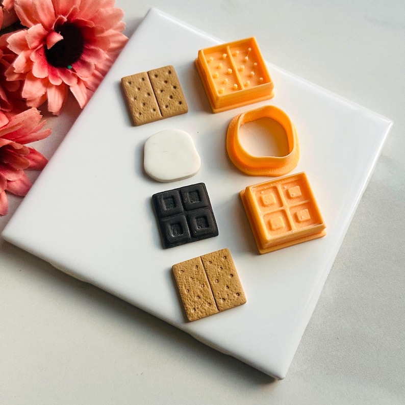 K&ouml;nnte beinhalten: Ein Set mit sieben Miniatur-Anh&auml;ngern aus Polymer Clay in Lebensmittelform, darunter zwei Graham-Cracker, ein Schokoriegel, ein Marshmallow und drei Waffel-Keksformen. Die Anh&auml;nger sind auf einer wei&szlig;en Oberfl&auml;che angeordnet.