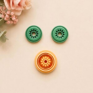May include: Two circular green buttons with a central gear-like cutout and one larger circular orange button featuring a detailed gear-shaped center. These three plastic buttons are arranged in a triangular formation on a light surface.