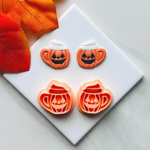 Puede incluir: Dos juegos de cortadores de galletas con temática de Halloween. El juego superior presenta tazas de calabaza naranja con crema batida blanca y caras de jack-o'-lantern negras. El juego inferior es el contorno del mismo diseño de taza de calabaza.