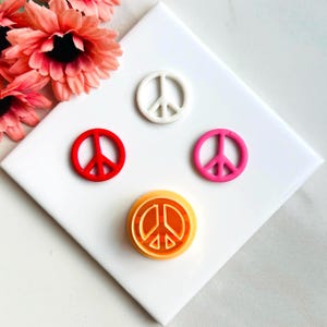 Può includere: Quattro tagliabiscotti a forma di segno di pace in diversi colori: bianco, rosso, rosa e arancione. I tagliabiscotti sono su una superficie bianca.
