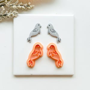 May include: Four seal-shaped cookie cutters and two seal-shaped decorations. The cookie cutters are orange, and the seals are grey with white dots. The items are arranged on a white square surface, with a sprig of white flowers in the upper left corner.