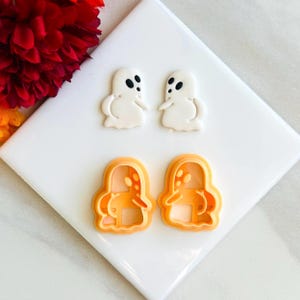May include: Two white ghost-shaped cookie cutters and two white ghost-shaped cookies on a white surface.