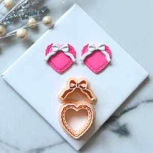 Può includere: Due orecchini a forma di cuore rosa con fiocchi bianchi e un tagliabiscotti a forma di cuore con un design a fiocco. Il tagliabiscotti è arancione chiaro e ha un bordo smerlato. Gli orecchini e il tagliabiscotti sono su una superficie bianca.