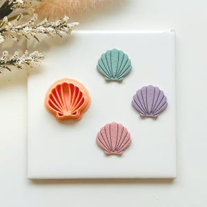 May include: A selection of seashell-shaped items in different colours. There is a coral-coloured seashell cookie cutter and three seashell-shaped decorations in teal, lavender, and pink. The items are arranged on a white surface.