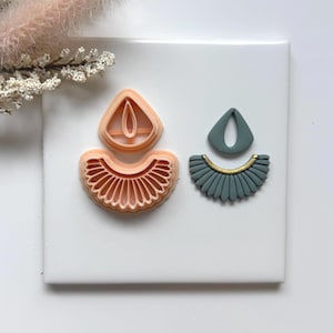 May include: A set of three earring-shaped clay cutters and a finished earring. The cutters are peach-colored plastic, and the finished earring is teal with a gold accent. The design features a teardrop shape and a fan-like bottom.