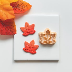 Puede incluir: Tres cortadores de galletas con forma de hoja de arce naranja y roja y dos galletas con forma de hoja sobre una superficie blanca. Las galletas tienen un degradado de rojo y naranja. El cortador de galletas es de color naranja claro. Hojas artificiales naranjas y rojas en el fondo.