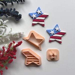 May include: A selection of star-shaped cookie cutters and star decorations. The decorations have a blue background with white stars and red and white stripes. The cookie cutters are peach-coloured and have the letter "M" on them.