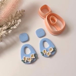 Può includere: Orecchini in pasta polimerica azzurra con dettagli floreali bianchi e borchie quadrate abbinate. Sono presenti anche taglierini in argilla color pesca, che suggeriscono un progetto fai-da-te. Gli orecchini hanno una forma a goccia.