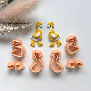 May include: An assortment of cookie cutters in the shape of geese and dresses. Two yellow and white goose-shaped cutters are at the top, with matching boots and hats. Below are several peach-colored dress-shaped cutters.