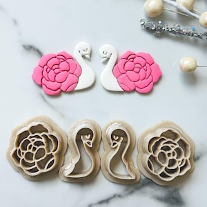 Puede incluir: Dos decoraciones de arcilla polimérica con forma de cisne y dos con forma de flor. Los cisnes son blancos con detalles rosas, y las flores son rosas. Debajo están los cortadores de galletas a juego en color marrón claro. Elementos decorativos para manualidades.