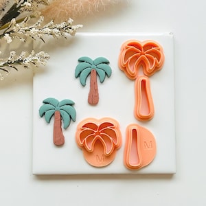 May include: A selection of palm tree-shaped cookie cutters in different sizes and styles. The cutters are in shades of orange and feature detailed palm fronds and trunks. Two palm trees are green and brown. The cutters are arranged on a white surface.