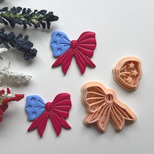 May include: Two red and blue bow-shaped cookie cutters with star details, and a bow-shaped cookie cutter. The bows have a blue section with white stars and a red section. The cookie cutters are on a white surface.