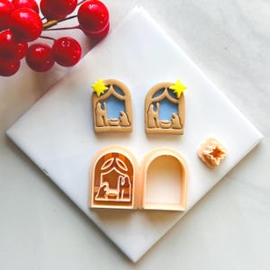 Puede incluir: Cortadores de galletas con forma de escena de la Natividad. Los cortadores son de color beige con un fondo azul y estrellas amarillas. La imagen también incluye un cortador en forma de estrella y bayas rojas.