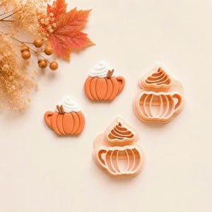 May include: Two orange pumpkin-shaped mug cookie cutters with white whipped cream tops and two finished orange pumpkin mug cookies with white whipped cream and brown cinnamon sticks, arranged with dried autumn leaves and berries.