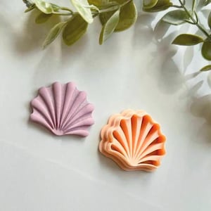 May include: Two seashell-shaped cookie cutters. One is a muted pink, the other is a light orange. Both have a detailed, ridged design. The cutters are on a white surface, with green leafy sprigs in the background.
