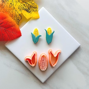 May include: A selection of corn-themed cookie cutters and decorations. Two corn-on-the-cob shapes with yellow kernels and teal husks are displayed above the word "YOU" spelled out with orange cookie cutters. Autumn leaves are in the background.