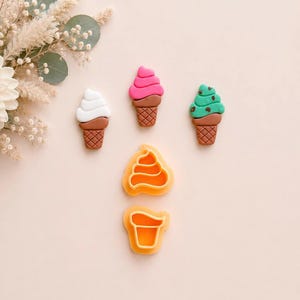 Könnte beinhalten: Drei kleine Eiswaffelfiguren in Weiß, Pink und Grün mit braunen Waffelstrukturen, begleitet von zwei orangefarbenen Kunststoff-Ausstechformen in Form eines Softeiswirbels und eines Waffelbodens auf einem hellen Hintergrund.
