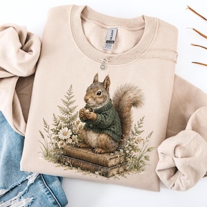 May include: Beige sweatshirt featuring a watercolor illustration of a squirrel wearing a green sweater, holding a nut, and sitting on books. The design includes flowers and ferns. The sweatshirt is paired with blue jeans.