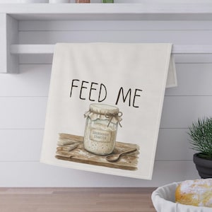 Puede incluir: Paño de cocina blanquecino con la frase "FEED ME" sobre una ilustración de un tarro de masa madre, una cuchara y espigas de trigo sobre una tabla de madera. El tarro tiene una etiqueta que dice "SOURDOUGH STARTER".