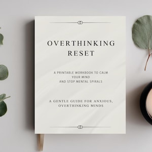 May include: A white workbook titled "OVERTHINKING RESET" with text, a lit candle in a black holder, and sprigs of greenery. The workbook is a guide for anxious minds.