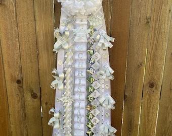 May include: White and silver graduation sash with the word "Senior" in silver lettering on a white flower. The sash has white and silver ribbons with the words "Class of 2023", "Christian", "Rising", "Stars", and "Senior" on them.