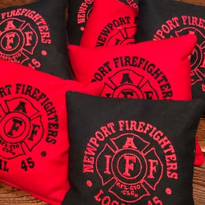 Può includere: Sacchi di fagioli rossi e neri con il testo "NEWPORT FIREFIGHTERS" e un emblema di pompiere con le lettere "IAFF" al centro. I sacchi di fagioli sono etichettati "LOCAL 45" e "AFL-CIO CLC."