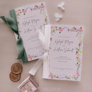 May include: Two wedding ceremony programs with floral borders and watercolor illustrations. The programs are tied together with a white ribbon and a green ribbon. The text on the programs reads "The Wedding Ceremony of Kyleigh Meyer and William Schmitt" and includes the date and time of the ceremony.