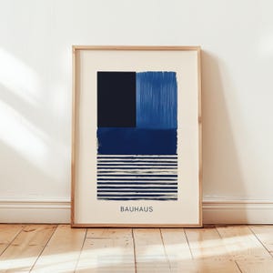 Könnte beinhalten: Ein gerahmter Druck mit einem geometrischen abstrakten Design in Blautönen. Der Druck zeigt ein dunkelblaues Quadrat, ein hellblaues Rechteck und ein dunkelblaues Rechteck mit weißen Streifen. Das Wort "BAUHAUS" ist in Schwarz am unteren Rand des Drucks gedruckt.