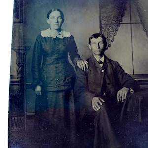 May include: A black and white tintype photograph of a couple posing for a portrait. The woman is wearing a dark dress with a white lace collar and the man is wearing a dark suit jacket. They are standing in front of a wall with a decorative pattern.