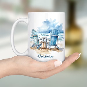 Peut inclure: Une tasse en c&eacute;ramique blanche avec une illustration aquarelle de deux chaises Adirondack bleues sur une plage avec un ciel nuageux et le nom "Barbara" imprim&eacute; au bas.