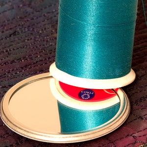 May include: A spool of teal thread with a white plastic bobbin and a red plastic base with the text "COV12" printed on it. The spool is sitting on a silver-colored, round, mirrored surface.